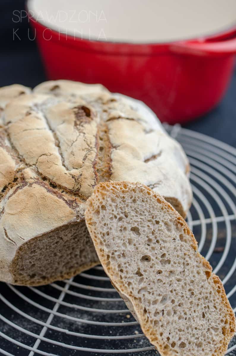 Bochenek chleba na kratce do studzenia z ukrojoną dużą kromką ukazującą ciemne wnętrze chleba.