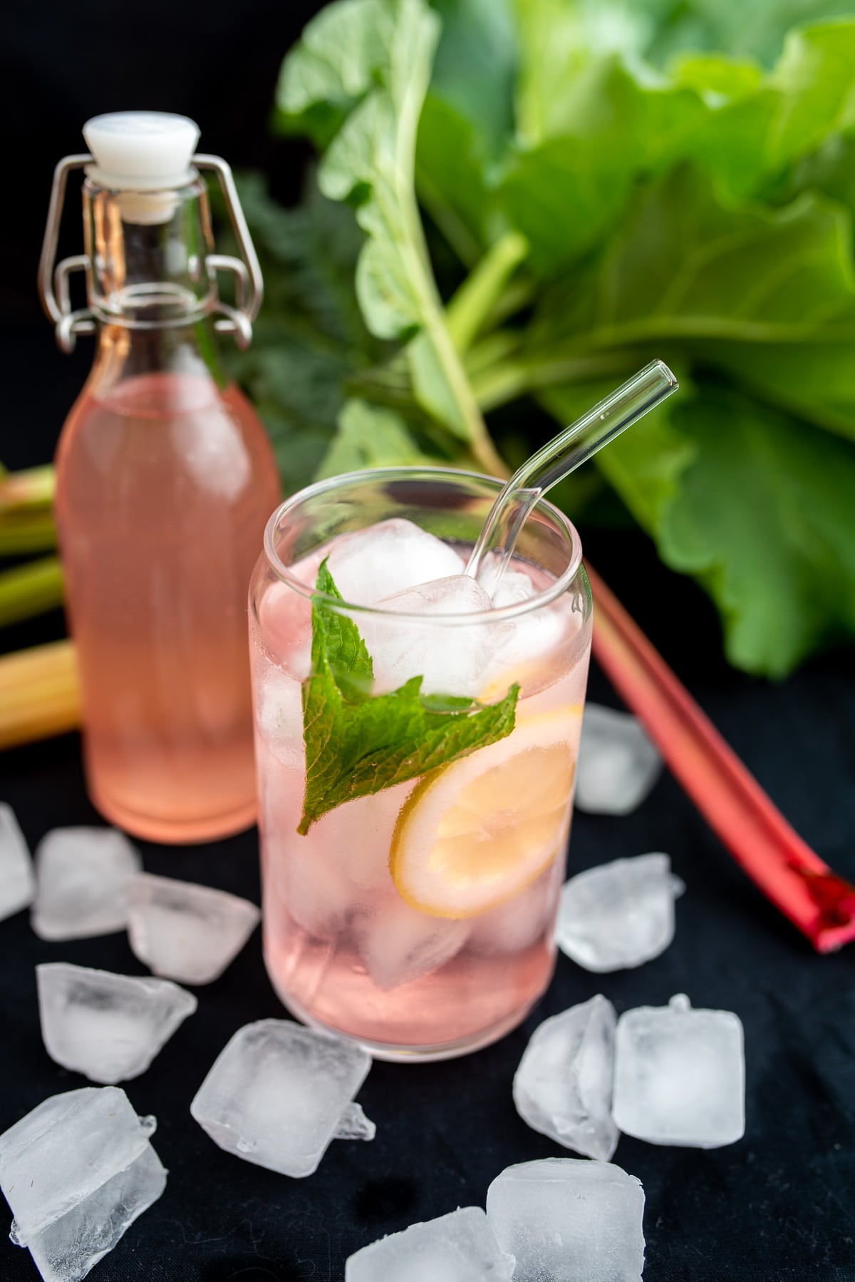 różowa lemoniad rabarbarowa w szklance pełnej lodu z cytryną i miętą w tle butelka z lemoniadą