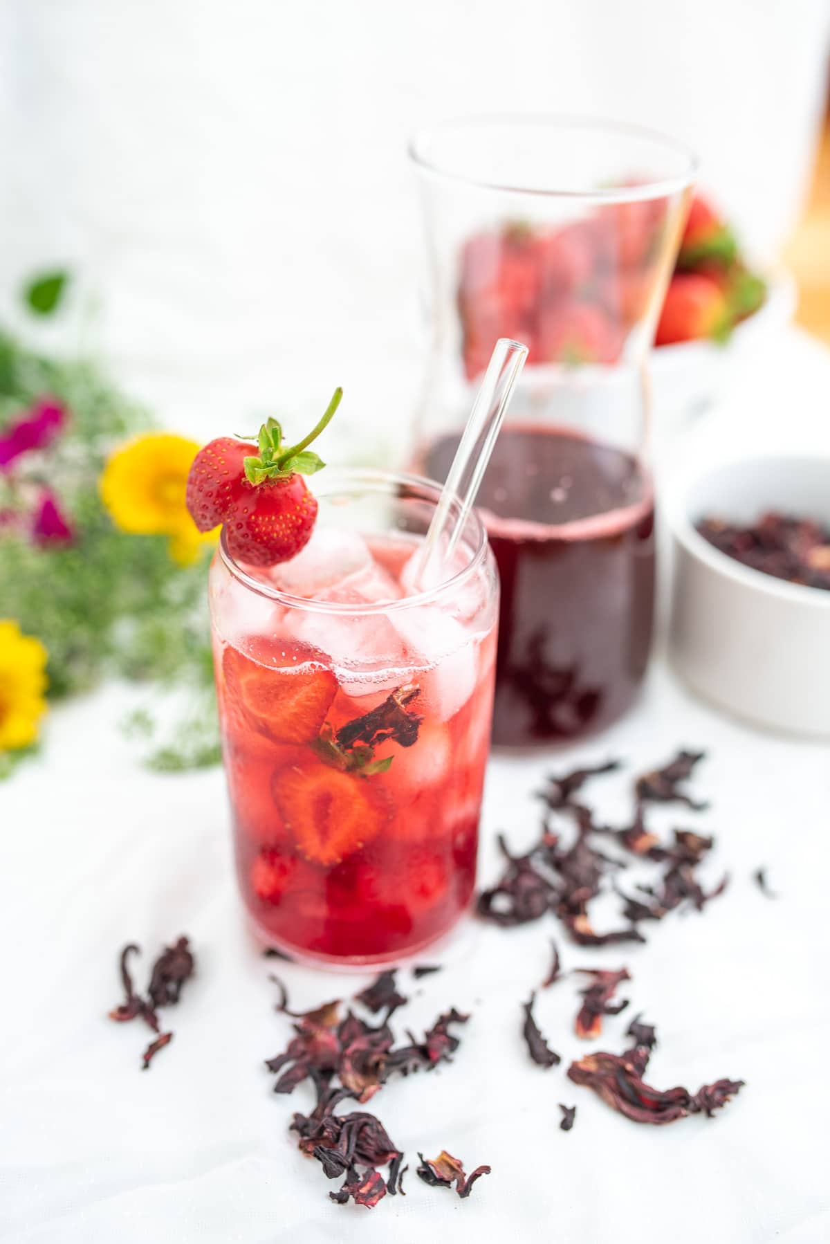 napój z hibiskusa w szklance wypełnionej wodą w tle butelka z napojem, truskawki i bukiet polnych kwiatów